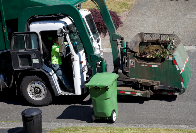 The cab and front loader of a garbage truck. The arm of the truck is about to empty a green organics tote into the front loader